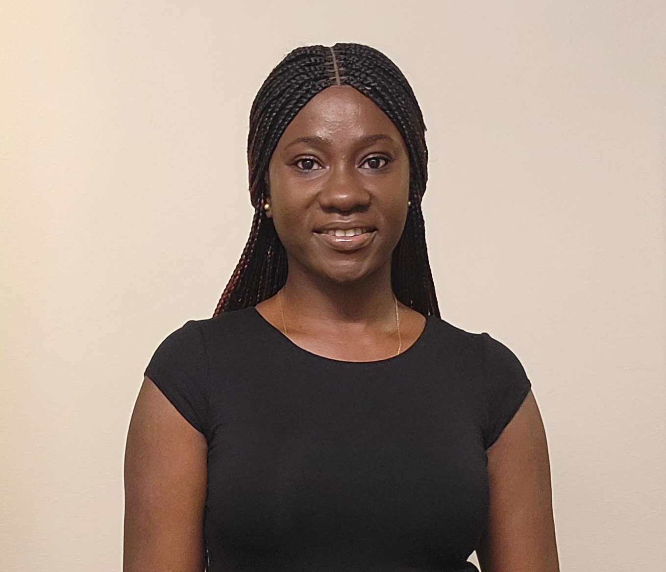 Professional photo of Oluwaseyi Aderinboye, a black woman with long black hair in braids. She is smiling and facing the camera, wearing a black t-shirt. Background is an off-white peachy color.